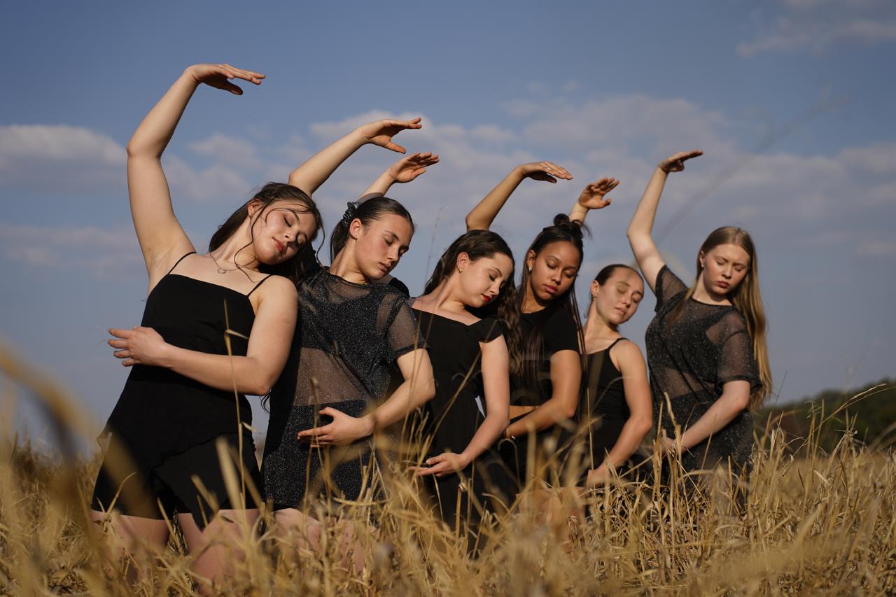 Six Diamond Dance dancers in black, arms raised, in a golden field